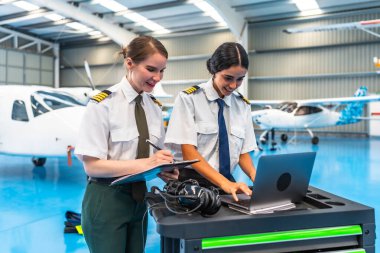 İki üniformalı kadın pilot, küçük uçaklarla çevrili geniş bir uçak hangarında dizüstü bilgisayar ve pano üzerinde çalışıyor.