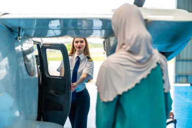 Müslüman bir yolcuyu küçük bir uçakta karşılayan kadın pilot, konukseverliği ve havacılığı somutlaştırıyor. Gülümseyen ve kapıyı açan, yolculuk için sıcak bir atmosfer yaratan