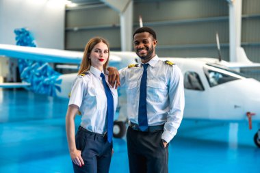 İki güler yüzlü havayolu pilotu özel bir jetin önünde, geniş bir hangarda, profesyonel yoldaşlıklarını sergileyerek güvenle poz veriyorlar.