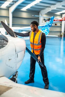 Yüksek görüşlü yelek giyen uçak bakım mühendisi hangardaki küçük bir uçağın pervanesini dikkatle inceliyor.