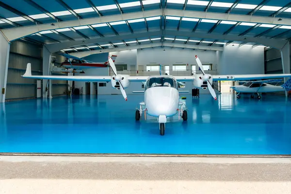 Geniş bir hangarın içine park etmiş beyaz çift motorlu uçak, parlak mavi bir zemine sahip ve etrafı daha küçük uçaklarla çevrili.