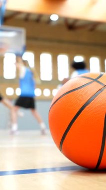 Turuncu basketbol topu, arka planda bayan basketbol oyuncularının antrenmanlarıyla parke sahada yatıyor.