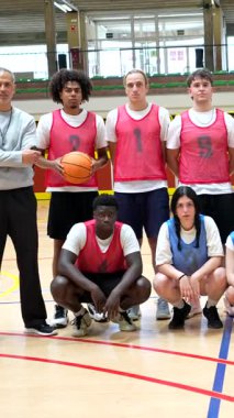 Numaralı formalar giyen iki basketbol takımı, kapalı sahada koçlarıyla fotoğraf çektirmek için poz veriyor.