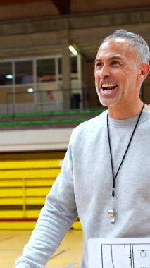 Deneyimli basketbol koçu, oyun stratejisini beyaz tahtayla açıklıyor ve oyuncularla spor salonunda tokalaşıyor.