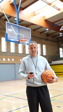 Top ve kronometre tutan gülümseyen bir basketbol koçunun portresi bir spor salonundaki basketbol sahasında duruyor.