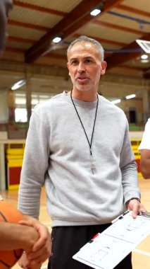 Basketbol koçu takıma yakın duruyor, stratejik oyun taktiklerini açıklamak için pano kullanıyor ve spor salonundaki antrenörlük tekniklerini kullanıyor.