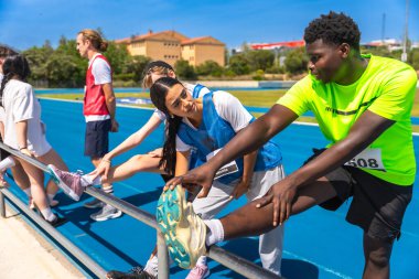 Atletizm sporcuları, güneşli bir günde yarışa hazırlanmak için pistin kenarındaki parmaklıklarda bacaklarını esnetiyorlar.