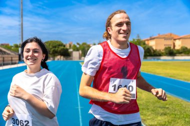 İki mutlu sporcu mavi gökyüzünün altında mavi bir pistte koşuyor.