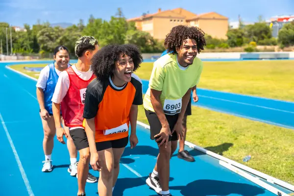 Bir grup sporcu, canlı mavi pistteki bir yarışı tamamladıktan sonra birlikte yaptıkları başarıları kutlayarak soluklanıp toparlanıyorlar.