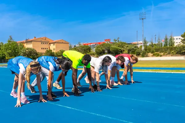 Çeşitli sporcu grupları mavi koşu pistinde yarışmaya hazırlar, yarışma ve atletizmi somutlaştırırlar.