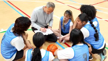 Basketbol koçu bir panoya oyun çiziyor ve liseli kızların basketbol takımına oyun stratejisini anlatıyor.