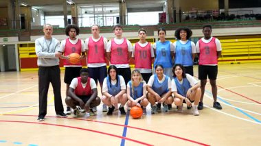 Lise basketbol takımı koçla bir araya geliyor, basketbol sahasında güven içinde poz veriyor, takım birliği ve atletik ruhu sergiliyor.