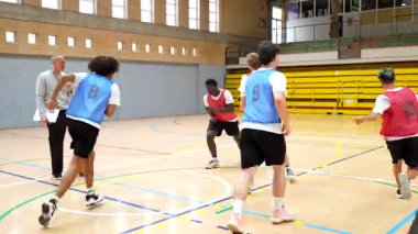 Genç basketbol oyuncuları antrenman sırasında kapalı bir sahada antrenman yaparken top sürme, pas verme ve savunma talimi yapıyorlar.