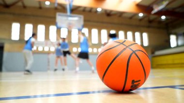Turuncu basketbol sahada dinleniyor. Dinamik antrenman sırasında arka planda bulanık oyuncular antrenman yapıyor.