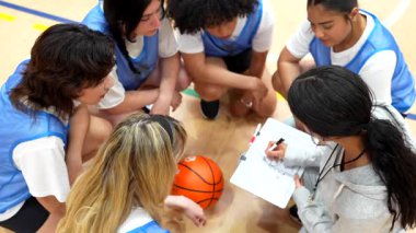 Basketbol koçu takım molası sırasında strateji belirliyor, spor salonundaki odaklanmış bayan sporcularla oyun taktiklerini konuşurken panoya resim çiziyor.