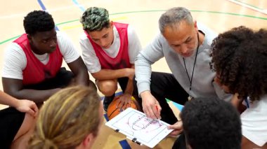 Basketbol koçu beyaz tahta ve işaretleyici kullanarak oyunculara stratejik oyun planlamasında rehberlik ediyor.