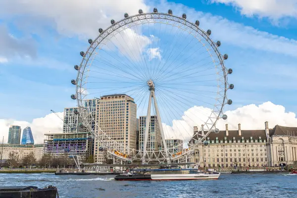 Londra 'nın gözü görkemli bir şekilde Thames nehrinin kıyısında dikiliyor, bir tekne geçiyor ve arkadaki modern binalar bulutlu mavi gökyüzünün altında.