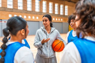 Kadın basketbol koçu top tutuyor ve spor salonunda antrenman yaparken takımına oyun stratejisini anlatıyor.