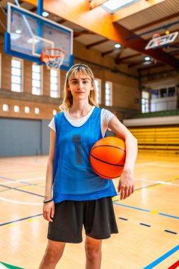 Genç bayan basketbolcu, elinde basketbol topuyla sahada duruyor, maç için hazır, özgüven ve kararlılık gösteriyor.