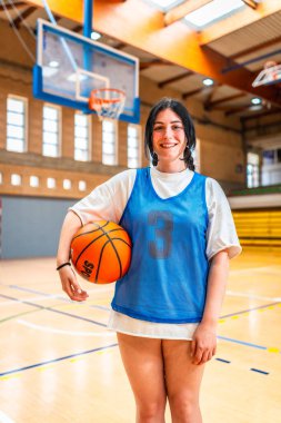 Spor salonunda gülümseyen ve elinde basketbol topu tutan bayan sporcu antrenman ya da yarışmaya hazır.