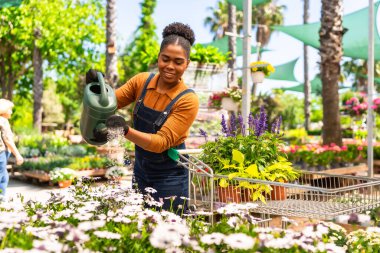 Bahçe merkezinde çalışan mutlu genç bir kadın bir alışveriş arabasını iterken bir su bidonuyla çiçekleri suluyor.