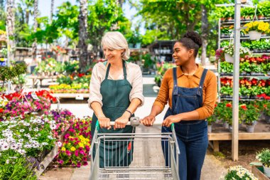 İki kadın bahçe çalışanı boş bir alışveriş arabasını renkli saksıların arasına itiyor.
