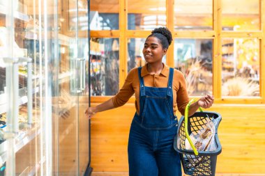 Gülümseyen siyah kadın marketin soğutmalı kapısını açıyor. Elinde alışveriş sepeti var.