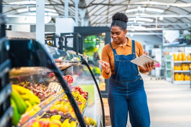 Genç bir süpermarket çalışanı dijital tablet kullanıyor ve soğutulmuş vitrindeki taze ürünleri kontrol ediyor.