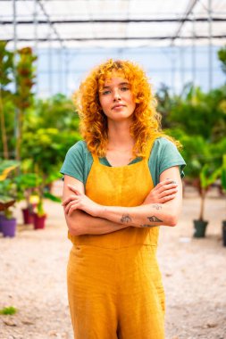 Kıvırcık kızıl saçlı ve dövmeli sarı tulumlu genç bir kadın. Çiçek serasında çapraz kollarıyla duruyor. Kendinden emin görünüyor.