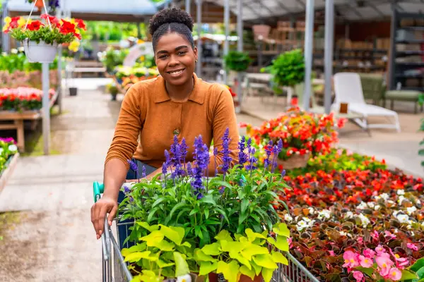 Gülümseyen Afro-Amerikalı bir kadın canlı çiçeklerle dolu bir alışveriş arabasını iterken hareketli bir bahçe merkezinde geziniyor.