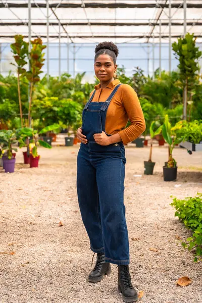 Kot pantolon giymiş, serada güvenle poz veren, bahçecilik uzmanlığını ve bitkilere olan tutkusunu sergileyen Afro-Amerikalı genç bir kadın.