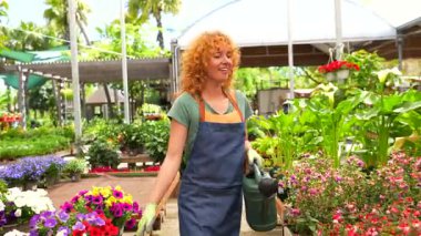 Kızıl saçlı bahçıvan kadın önlük ve eldiven giyer, serada çalışırken su bidonu tutar, çiçek ve bitkilerle ilgilenir.