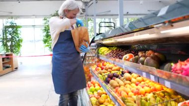Üst düzey kadın manav, yerel bir markette çalışırken olgun, renkli meyveleri dikkatlice seçiyor ve kağıt torbaları dolduruyor.