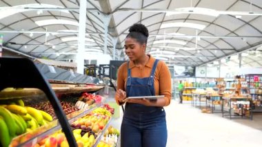 Genç Afro-Amerikan süpermarket çalışanı markette taze meyve kontrolü için dijital tablet kullanıyor.