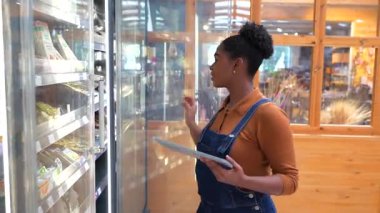Modern marketin iç kısımlarında soğutulmuş rafların yanında dururken dijital tablet kullanarak envanter taraması yapan profesyonel kadın perakendeciler.