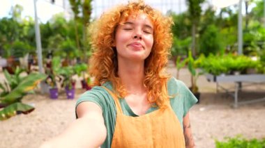 Bahçıvan olarak çalışan genç kızıl saçlı kadın kameraya el ele tutuşuyor, gülümsüyor ve serada onu takip etmeye davet ediyor.