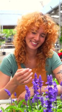 Kızıl saçlı kadın bahçıvan, seranın içinde mor çiçekleri besliyor, neşeli gülümseme ve profesyonel bakımla bitkilerle ilgileniyor.