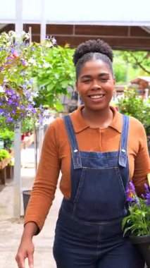 Mor çiçek sepeti taşırken gülümseyen kadın bahçıvan, neşeli girişimci ruhuyla bahçe merkezinde yürüyor, küçük işletme yaşam tarzını gözler önüne seriyor.