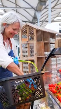 Kıdemli kadın manav dikkatlice taze, renkli ürünleri seçerken sıcak bir gülümseme ve yerel pazar ortamında müşterilere hizmet ediyor.