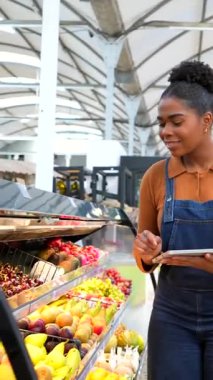 Genç Afrikalı Amerikalı satıcı dijital tablete not alıyor ve süpermarkette taze ürünleri kontrol ediyor.