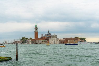 San Giorgio Maggiore kilisesi ve ikonik çan kulesinin manzarası, bulutlu bir günde sakin gölde zarif bir şekilde yüzen tekneler.