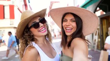 Güneş şapkalı iki genç kadın hediyelik eşya dükkanlarıyla dolu bir caddede yürürken selfie çekiyorlar.