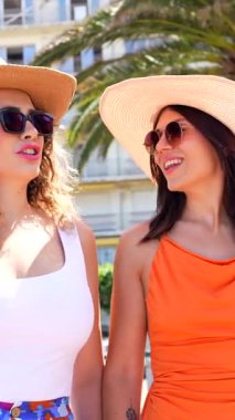 Two happy women wearing summer clothes, sunglasses and straw hats are walking hand in hand and chatting on a sunny day