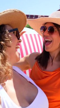 Şık mayo ve güneş şapkalı genç bayan arkadaşlar güneşli kumsal anlarının tadını çıkarıyorlar, neşeli selfie ve tasasız pozlarla tatil anılarını yakalıyorlar.