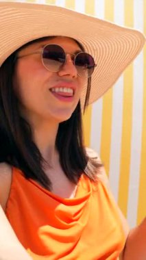 Mutlu bir turist çizgili bir plaj sandalyesinde güneş gözlüğü ve turuncu bir bluzla yaz tatilinin tadını çıkarıyor.