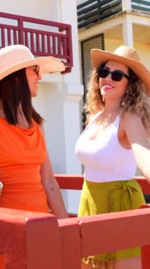 Güneş gözlüğü takan ve yaz şapkası takan iki neşeli kadın turist güzel bir sahil kasabasında yürüyüp tatillerinin tadını çıkarıyorlar.