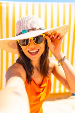 Genç bir kadın güneşli bir sahilde selfie çekiyor, turuncu bir elbise ve geniş kenarlı güneş şapkası giyiyor.