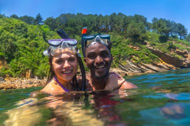 Açık mavi denizde şnorkelle yüzen ırklar arası bir çift, çarpıcı yeşil bir uçurumla çevrili, birlikte neşeli bir yaz macerasının tadını çıkarıyorlar.