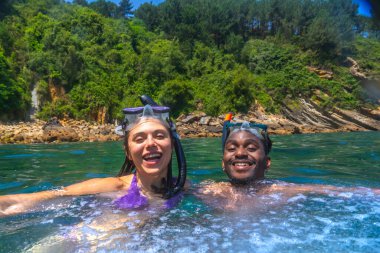 Irklar arası çift, arka planda yemyeşil bitki örtüsü olan güzel turkuaz sularda şnorkelle yüzmeyi seviyor.