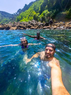 Üç arkadaş şnorkel kıyafeti giyip kayalık bir sahilin yakınındaki ferahlatıcı okyanus suyunun tadını çıkarıyorlar.